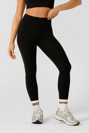 A person in a black moisture-wicking sports bra, white socks, silver running shoes, and Lorna Jane Lotus No Chafe Ankle Biter Leggings stands against a plain light grey background. Their head is not visible.