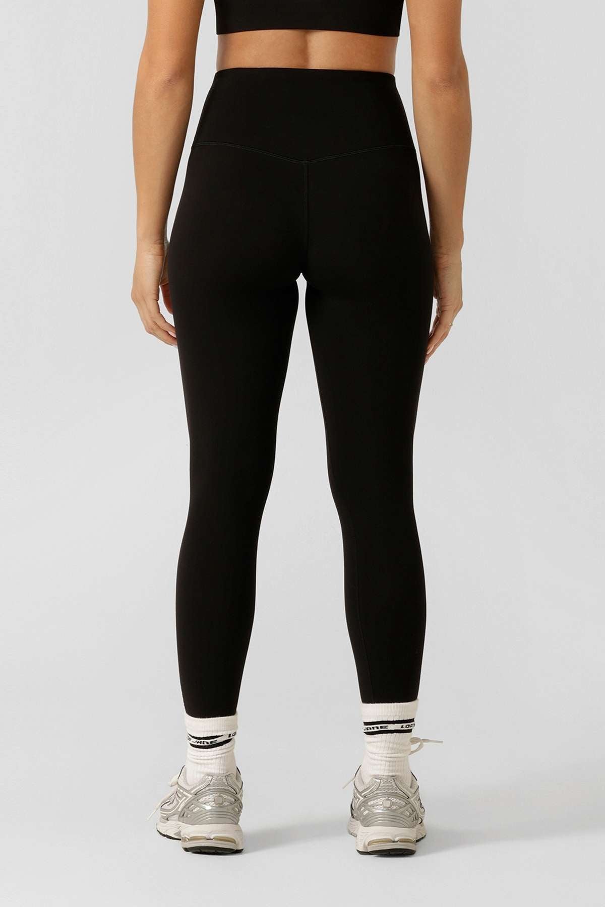 A woman faces away, wearing a black sports bra and Lorna Jane Lotus No Chafe Ankle Biter Leggings made from 4-way stretch, moisture-wicking fabric, plus white socks and gray athletic shoes against a light gray background.