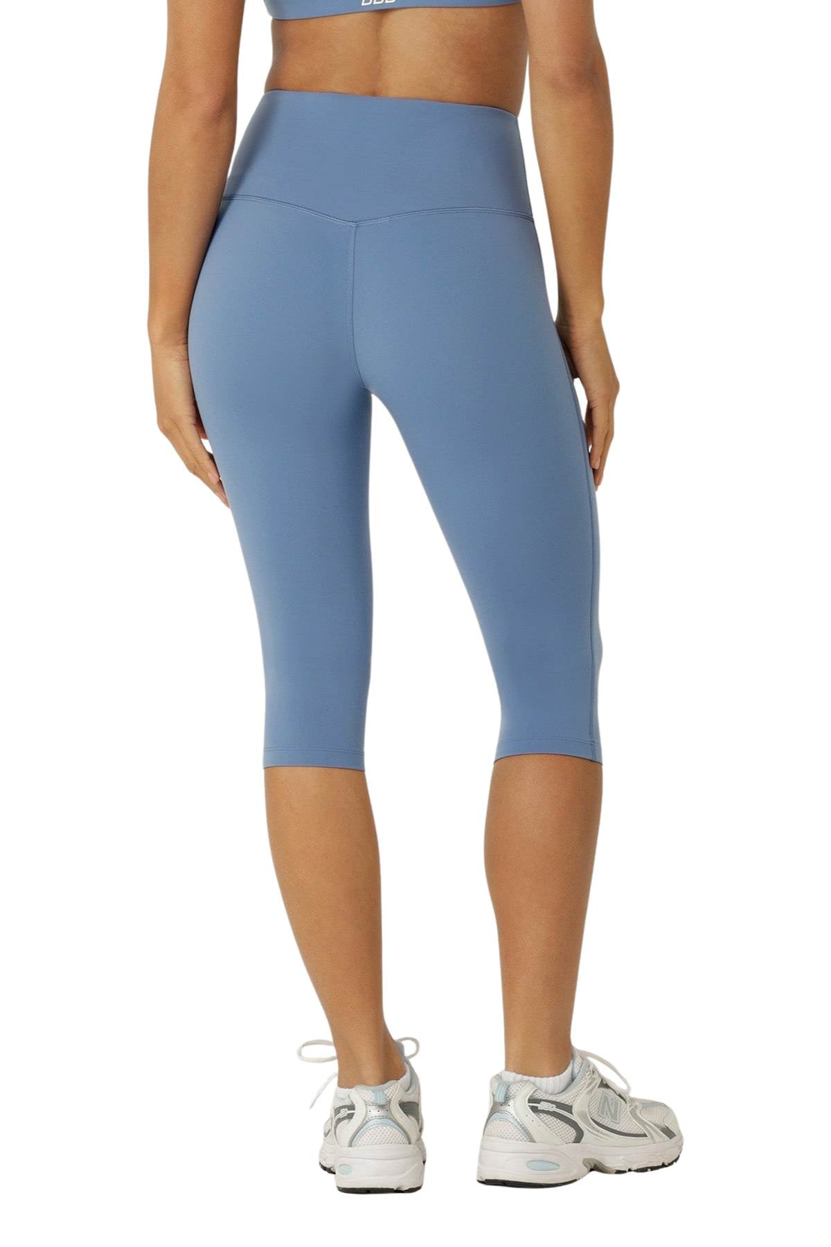 Woman wearing light blue Lorna Jane Lotus No Chafe Phone Pocket 3/4 Leggings and white sneakers, standing with her back to the camera against a white background.