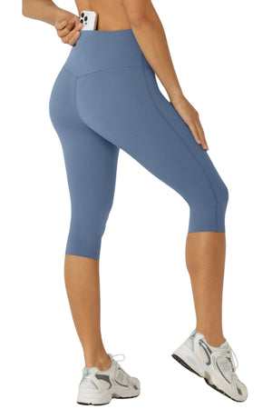 A person wearing Lorna Jane Lotus No Chafe Phone Pocket 3/4 Leggings in blue places a smartphone into the back pocket, showing off the stylish, moisture-wicking fabric and knee-length design. Only their lower body, paired with white sneakers, is visible.