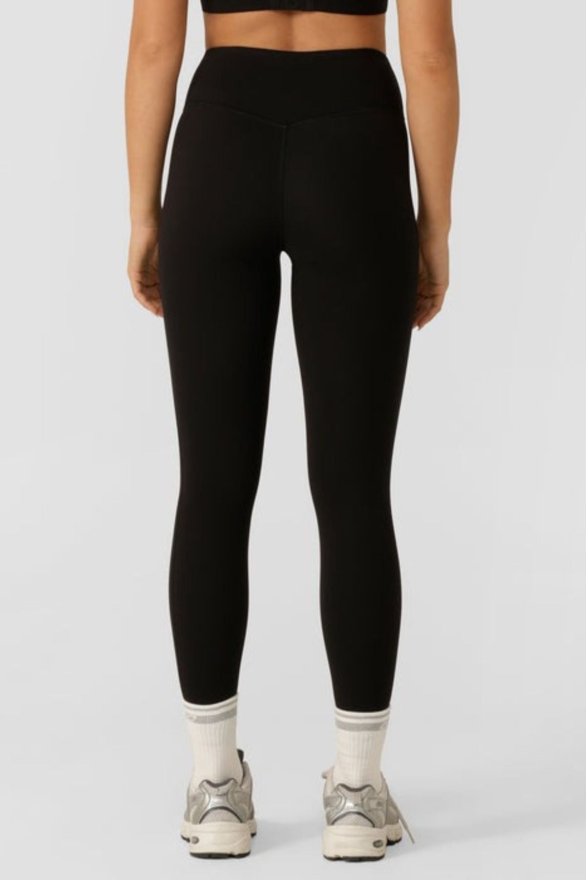 A person shown from the back against a plain light gray background wears black Lorna Jane Lotus No Chafe Phone Pocket Ankle Biter Leggings, white socks, and white athletic shoes.