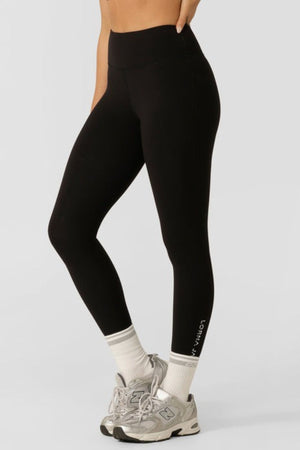 A person wearing Lorna Jane Lotus No Chafe Phone Pocket Ankle Biter Leggings, white crew socks, and gray athletic sneakers stands against a plain light background. The leggings feature subtle lower leg text.