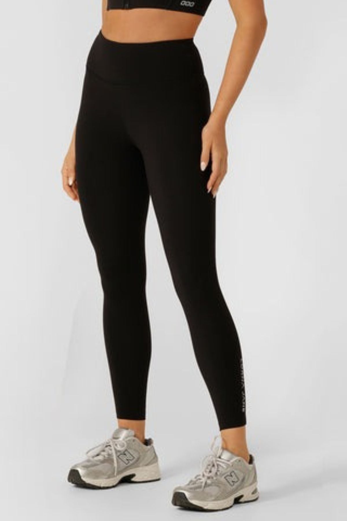 A person’s lower half is shown, wearing Lorna Jane Lotus No Chafe Phone Pocket Ankle Biter Leggings in black, paired with a black crop top and gray New Balance sneakers, standing against a light background highlighting their activewear.