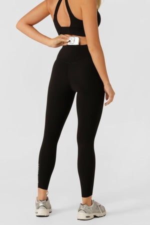 A woman in black activewear faces away, holding a white card at the waist of her Lorna Jane Lotus No Chafe Phone Pocket Ankle Biter Leggings. She also wears a black sports bra and gray sneakers against a plain light background.