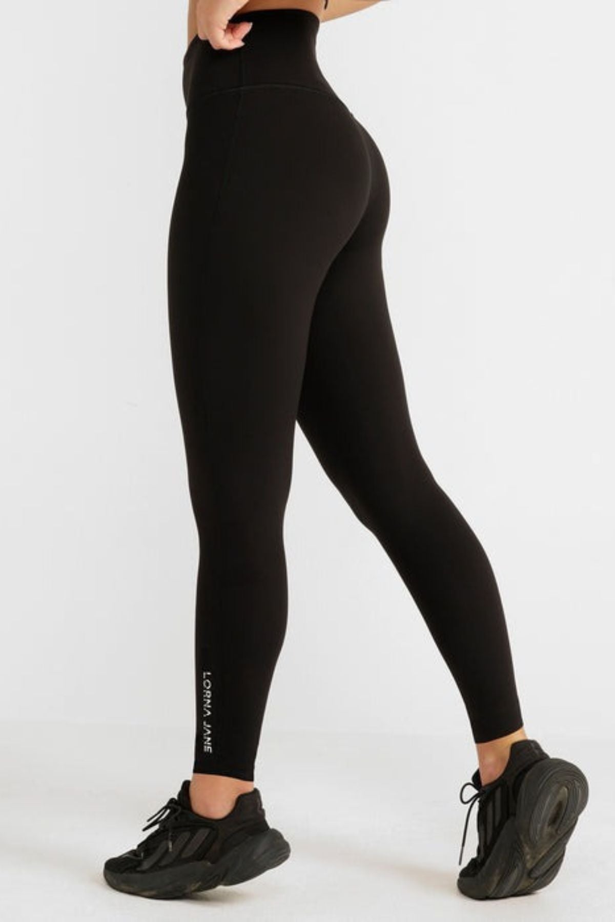 A person wearing black Lorna Jane Lotus No Chafe Full Length Leggings and black sneakers stands against a white background. The leggings, offering friction-free movement, display "Lorna Jane" in small white letters near the left ankle.