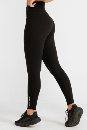 A person wearing black Lorna Jane Lotus No Chafe Full Length Leggings and black sneakers stands against a white background. The leggings, offering friction-free movement, display "Lorna Jane" in small white letters near the left ankle.