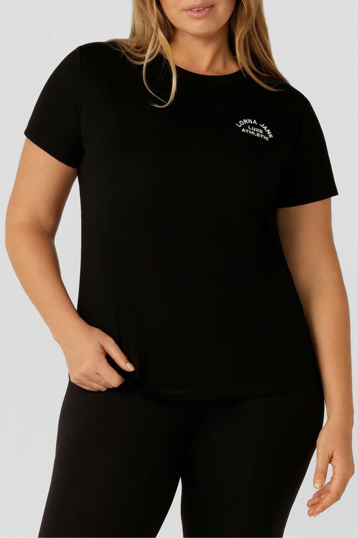 A woman wears the Lorna Jane Lotus T-Shirt in black, featuring a small white "Lorna Jane Always Active" logo on the upper left chest, paired with black pants—an ideal outfit for an active lifestyle.