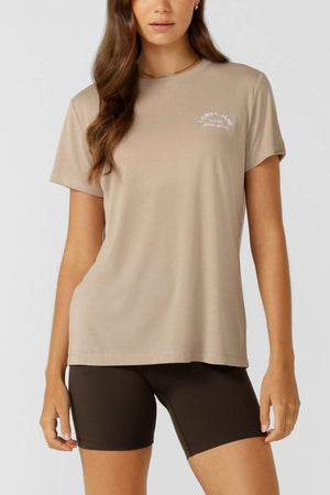 A woman wears a beige Lorna Jane Lotus T-Shirt with an embroidered logo on the left chest and black fitted shorts, standing facing forward with one arm relaxed by her side against a plain light gray background.