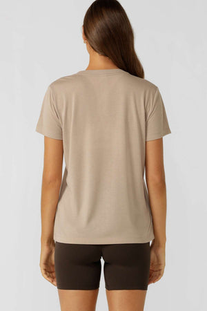 A woman faces away from the camera in a beige Lorna Jane Lotus T-Shirt with an embroidered logo and dark brown shorts, set against a light backdrop—ideal for an active lifestyle.