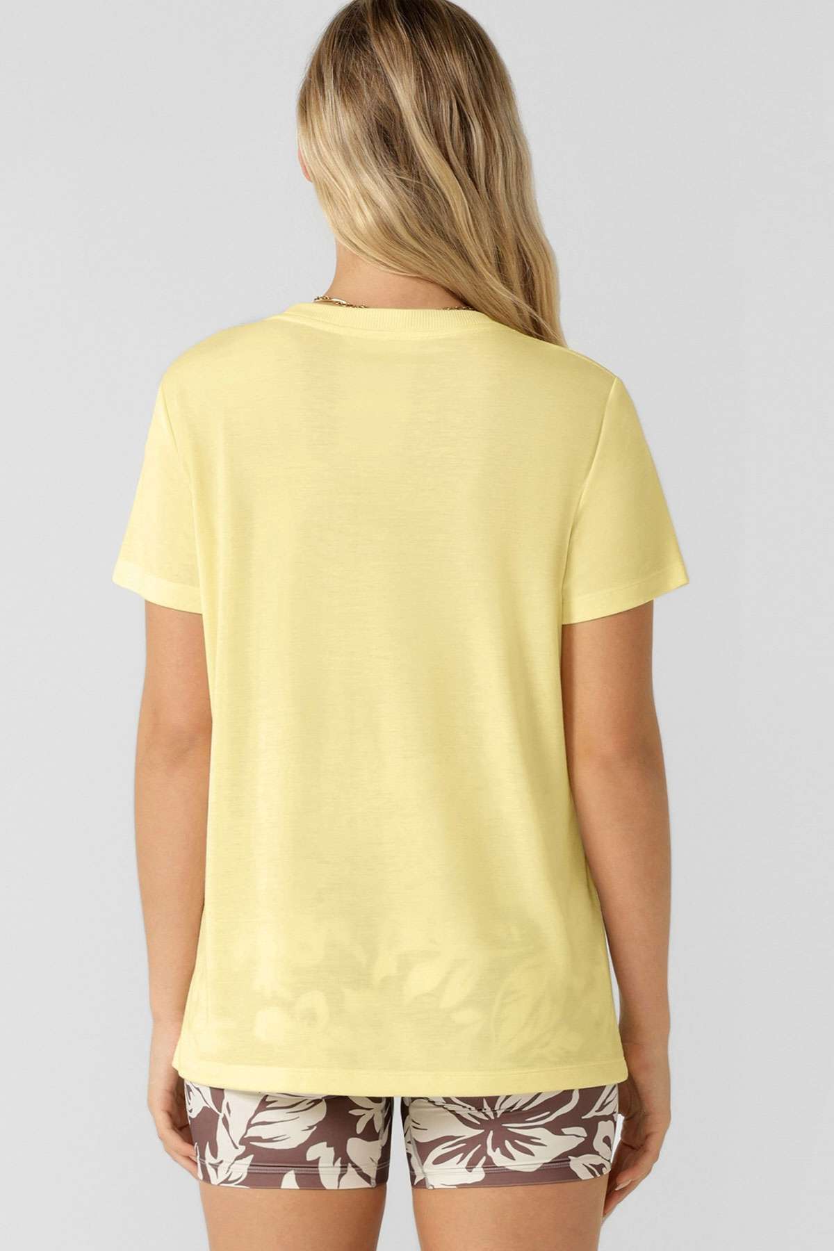 A woman with long blonde hair is seen from behind, wearing a light yellow Lorna Jane Lotus T-Shirt and brown shorts with a white floral pattern, standing against a plain light gray background.