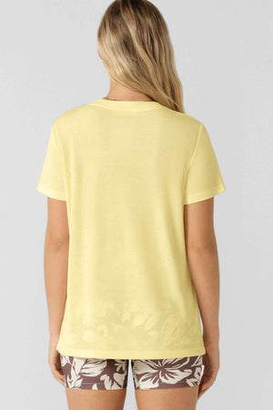 A woman with long blonde hair is seen from behind, wearing a light yellow Lorna Jane Lotus T-Shirt and brown shorts with a white floral pattern, standing against a plain light gray background.