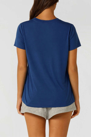 Shown from behind, a person with long brown hair wears a blue Lorna Jane Lotus T-Shirt with an embroidered logo and light gray shorts against a plain background—ideal for an active lifestyle.