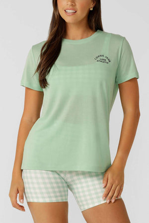 A woman stands against a plain background wearing a light green Lorna Jane Lotus T-Shirt with chest text and matching green and white gingham shorts—an ideal outfit for an active lifestyle.