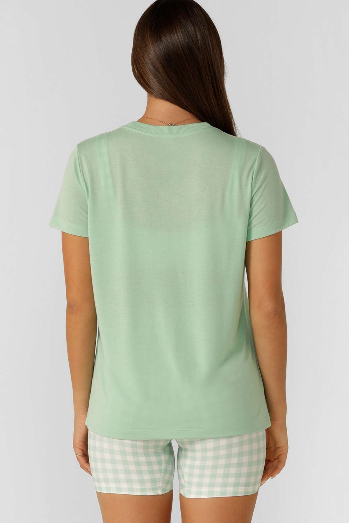 A woman with long brown hair faces away, wearing the Lorna Jane Lotus T-Shirt and pastel green-and-white checkered shorts—ideal for an active lifestyle—against a plain light gray background.