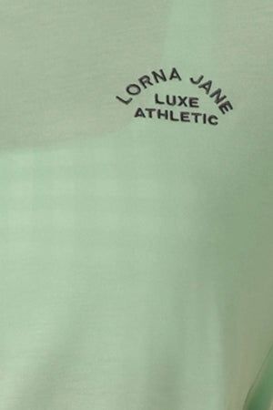 The Lorna Jane Lotus T-Shirt features light green fabric, black curved "LORNA JANE LUXE ATHLETIC" text on the top left, and 2-way stretch for comfort during your active lifestyle.