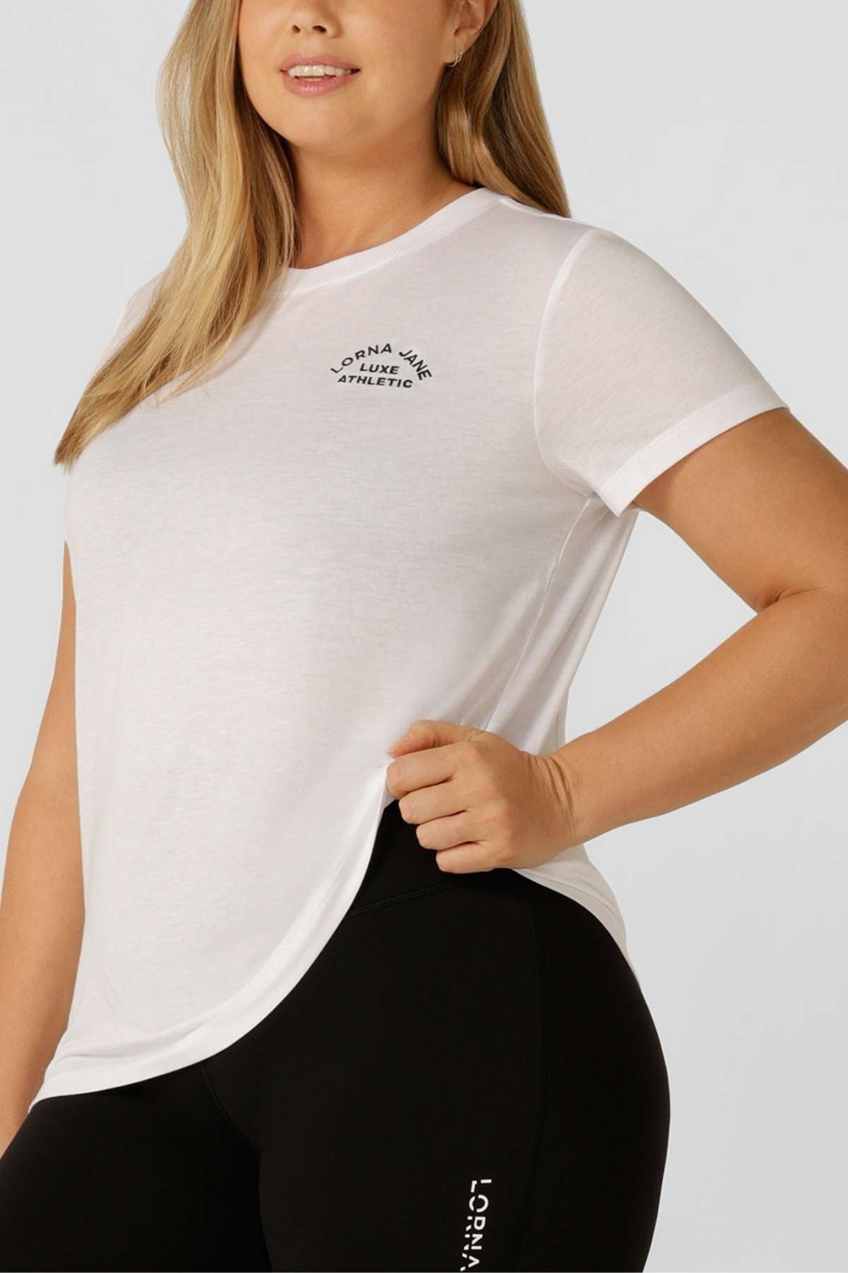 A woman wears a fitted white Lorna Jane Lotus T-Shirt with an embroidered chest logo and black leggings featuring white side text, standing against a plain light background.