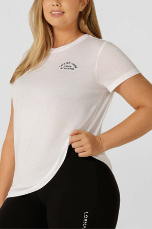 A woman wears a fitted white Lorna Jane Lotus T-Shirt with an embroidered chest logo and black leggings featuring white side text, standing against a plain light background.