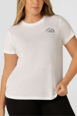 A woman wears the Lorna Jane Lotus T-Shirt with a small "Lorna Jane Athletic" logo on the left chest, pairing it with black pants against a light gray background—ideal for an active lifestyle.