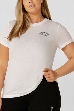 A woman wears the fitted Lorna Jane Lotus T-Shirt in white, featuring an embroidered logo, paired with black leggings. She holds the hem of her shirt, reflecting an active lifestyle.