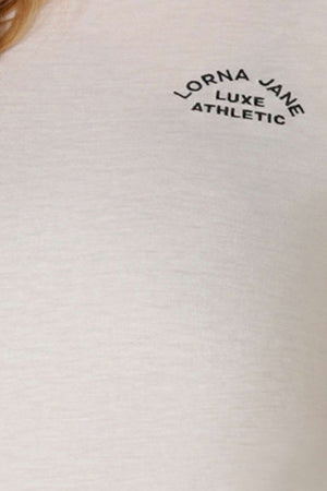 Close-up of a light-colored Lorna Jane Lotus T-Shirt featuring "LORNA JANE LUXE ATHLETIC" in black above the left chest—ideal for an active lifestyle.