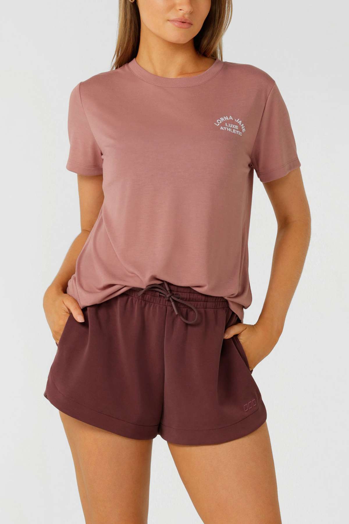 A woman wears a mauve Lorna Jane Lotus T-Shirt with 2-way stretch, tucked into high-waisted maroon drawstring shorts, standing hands-in-pockets against a plain light background—perfect for an active lifestyle.