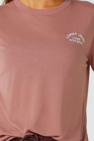 A person wears the mauve-pink Lorna Jane Lotus T-Shirt featuring white "Lorna Jane Luxe Athletic" text. The cropped image focuses on the upper torso, showcasing the 2-way stretch fabric—ideal for an active lifestyle.