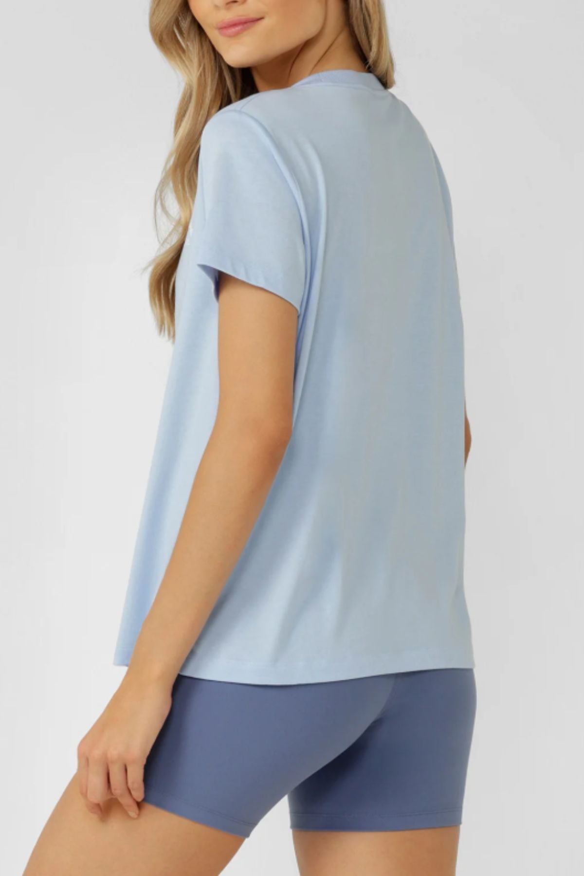 A woman with long blonde hair models the Lorna Jane Lotus T-Shirt and medium blue fitted shorts, standing sideways and facing away from the camera against a plain light background, exuding an active lifestyle.
