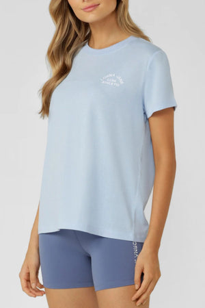 A woman in a light blue Lorna Jane Lotus T-Shirt and fitted blue shorts, both featuring subtle logo text, stands before a plain background. The long line silhouette of her outfit highlights an active lifestyle; her face is partially out of frame.
