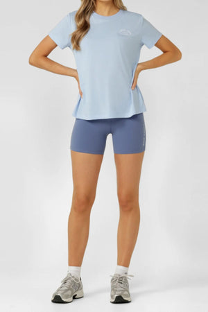 A woman stands with hands on hips, dressed in a Lorna Jane Lotus T-Shirt and blue athletic shorts. She wears white socks and gray sneakers against a plain light background. Her head is partially out of frame, highlighting the active outfit.