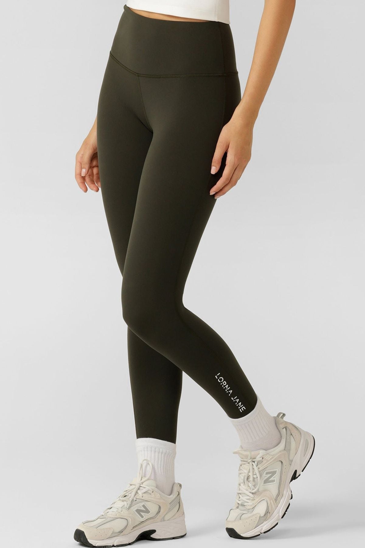 Someone stands against a plain light background wearing dark green Lorna Jane Lotus No Chafe Phone Pocket Ankle Biter Leggings, white socks, and New Balance sneakers. "Lorna Jane" appears near the left ankle on the activewear.