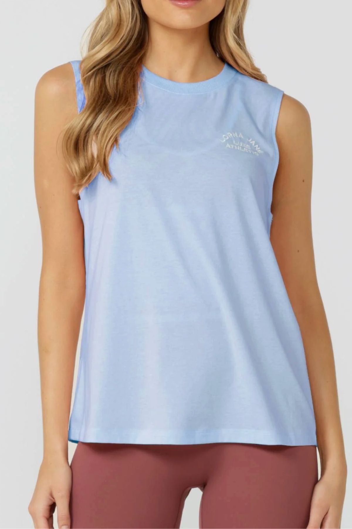 A woman stands forward wearing the Lorna Jane Lotus Muscle Tank in light blue with mauve leggings. The breathable tank has a round neckline and subtle white chest text. Her hair falls in loose waves.