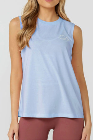 A woman stands forward wearing the Lorna Jane Lotus Muscle Tank in light blue with mauve leggings. The breathable tank has a round neckline and subtle white chest text. Her hair falls in loose waves.