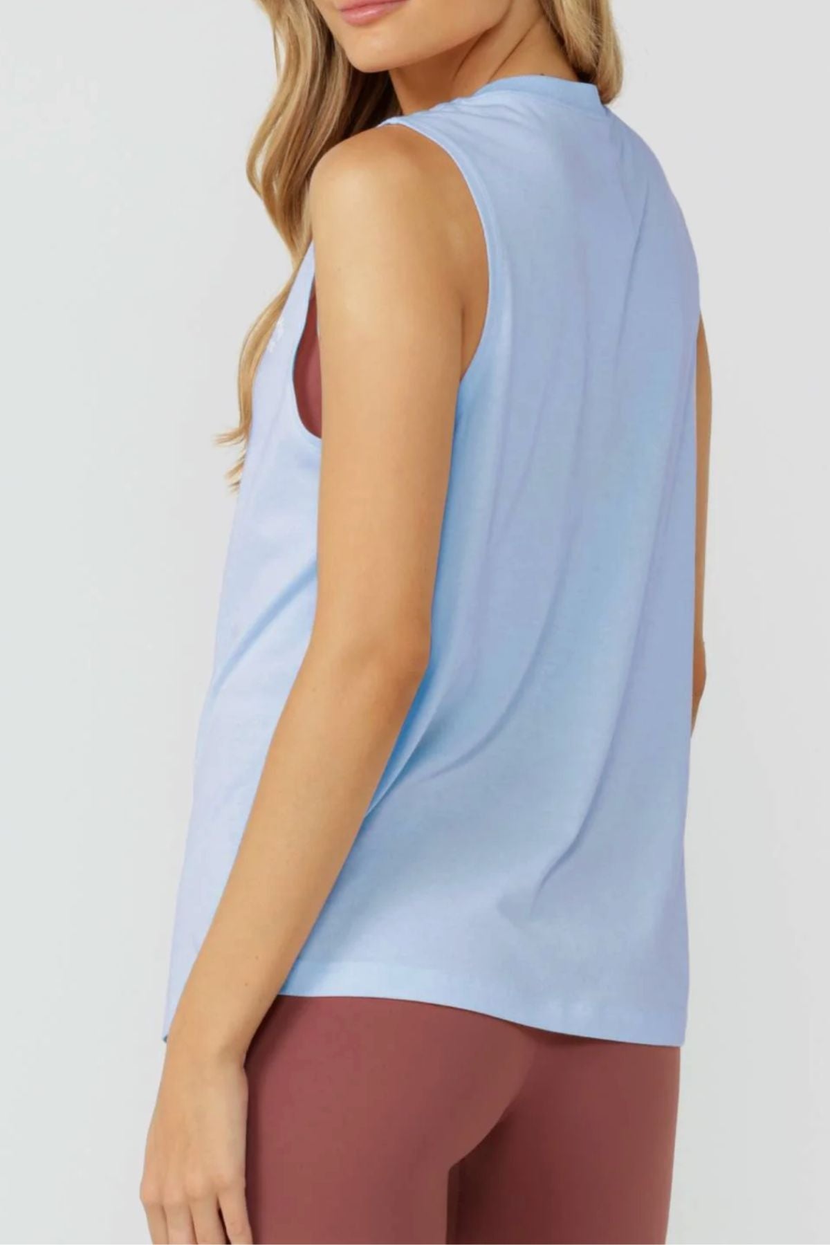 A woman with long blonde hair wears the Lorna Jane Lotus Muscle Tank over a maroon sports bra and leggings, posing with her back slightly turned to the camera against a light background.