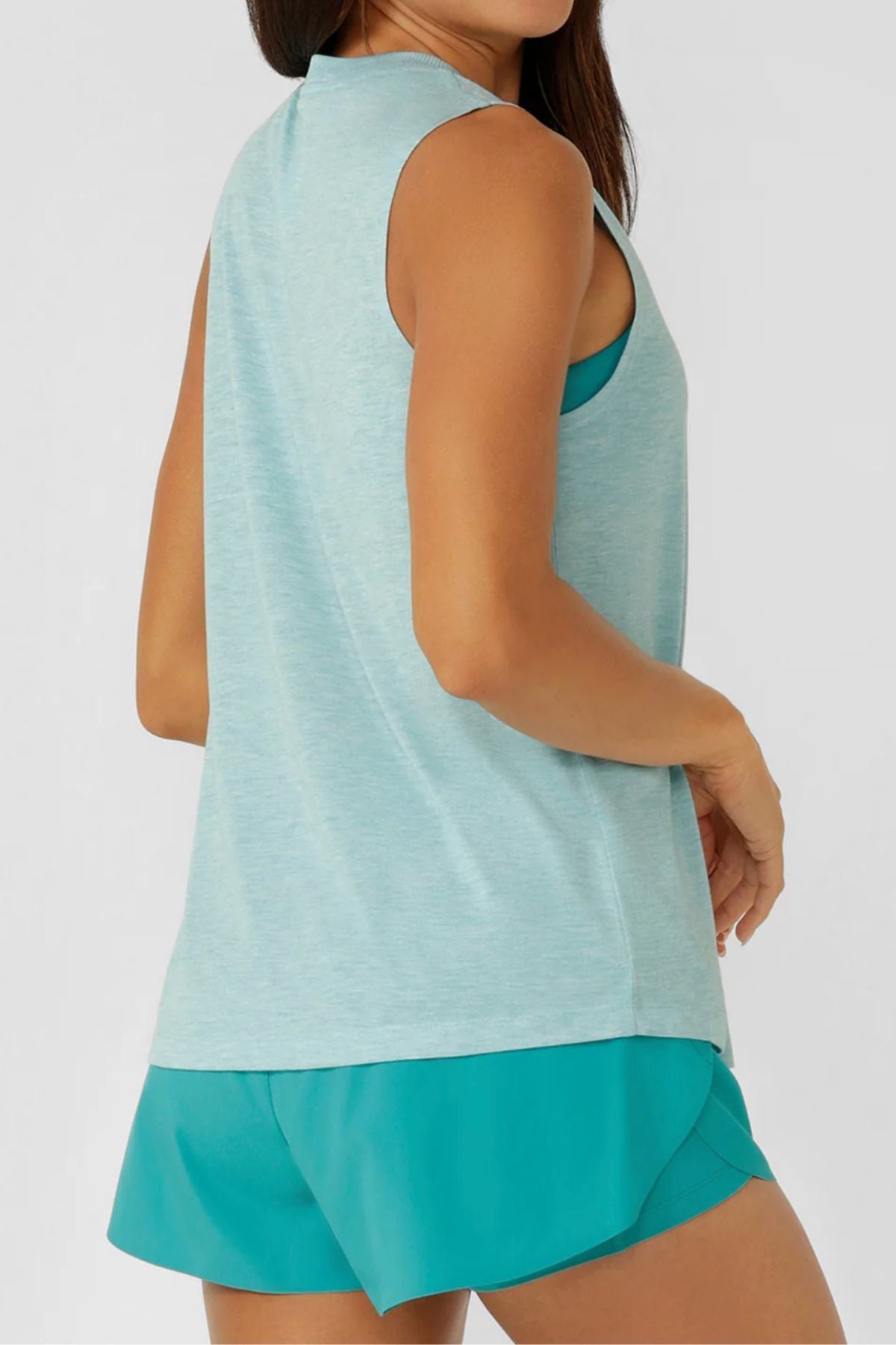 A woman models the Lorna Jane Lotus Muscle Tank in light blue with teal shorts, shown from side and back angles. The relaxed, long-line silhouette stands out against a plain light background.