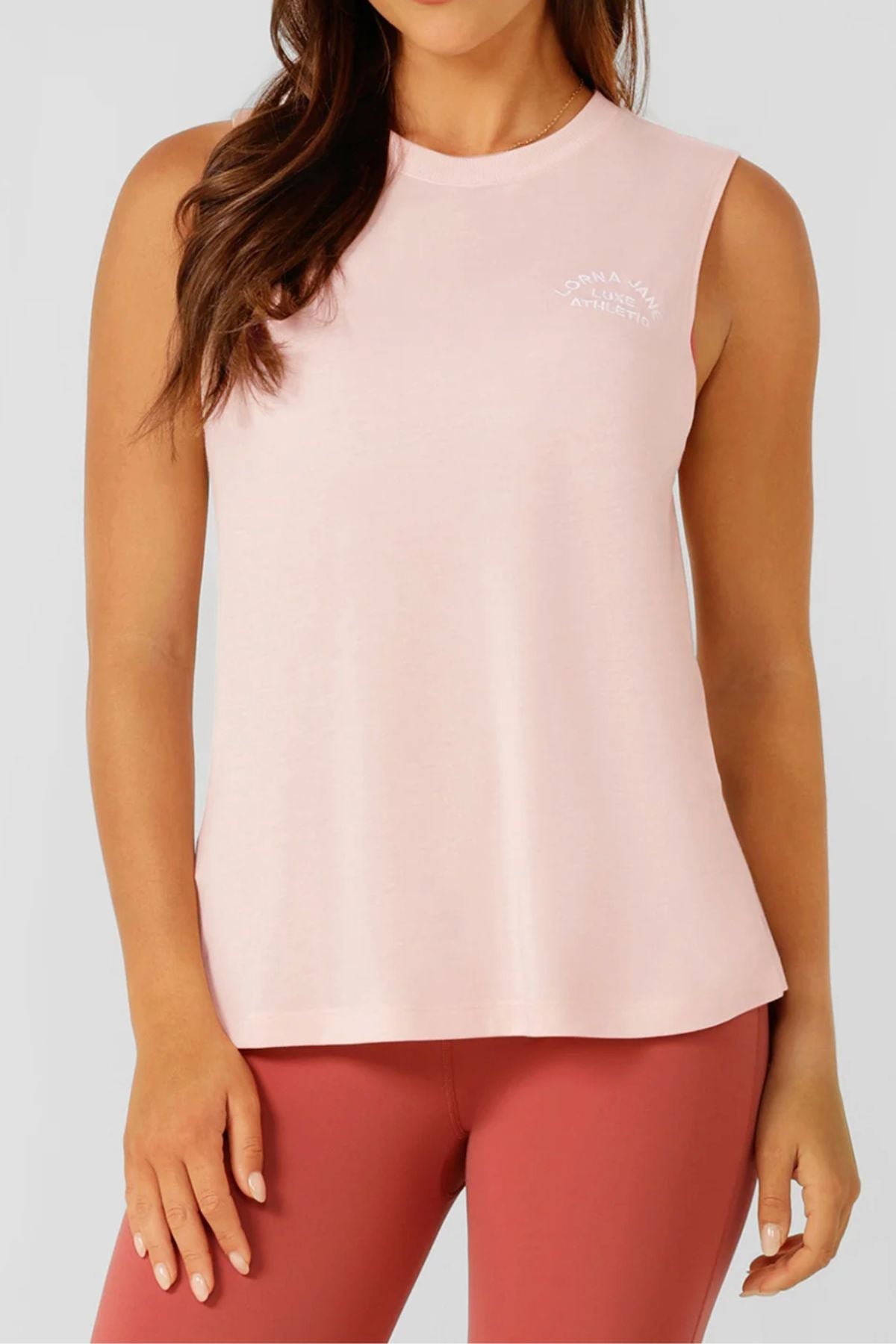 A person wearing the Lorna Jane Lotus Muscle Tank in light pink with white chest text, paired with high-waisted mauve leggings, stands against a plain light gray background.