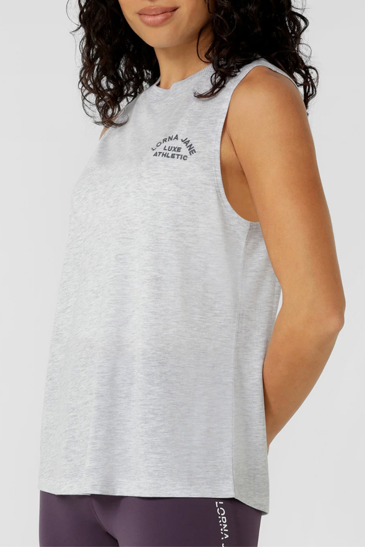A woman models the Lorna Jane Lotus Muscle Tank in light gray, featuring "Lorna Jane Luxe Athletic" on the chest, paired with purple leggings. She poses to highlight its lightweight, breathable natural blend fabric.