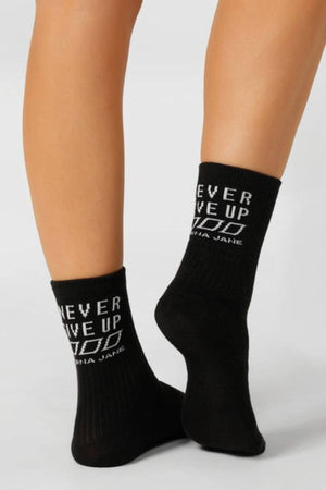 A person stands on their toes with legs slightly crossed, wearing Lorna Jane Never Give Up Arch Support Crew Socks—black with white "NEVER GIVE UP" text and a small design. These socks combine style and comfort on a light background.