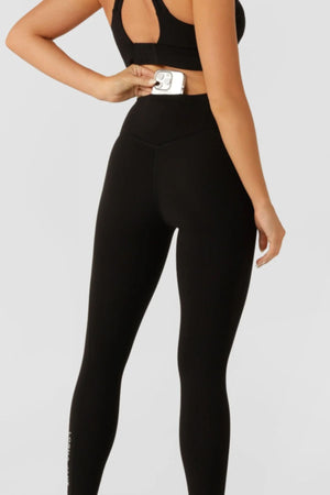 A woman in black Lorna Jane Lotus No Chafe Phone Pocket Full Length Leggings, seen from behind, has one arm bent and the other relaxed at her side.