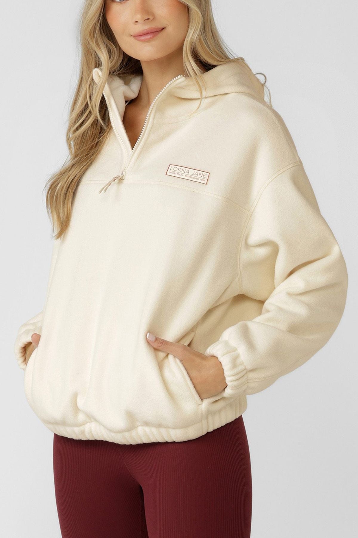 A person stands against a plain light background wearing the cream-colored Lorna Jane Nova Polar Fleece Quarter Zip Hoodie with a hood and front pockets, paired with burgundy Lorna Jane leggings.