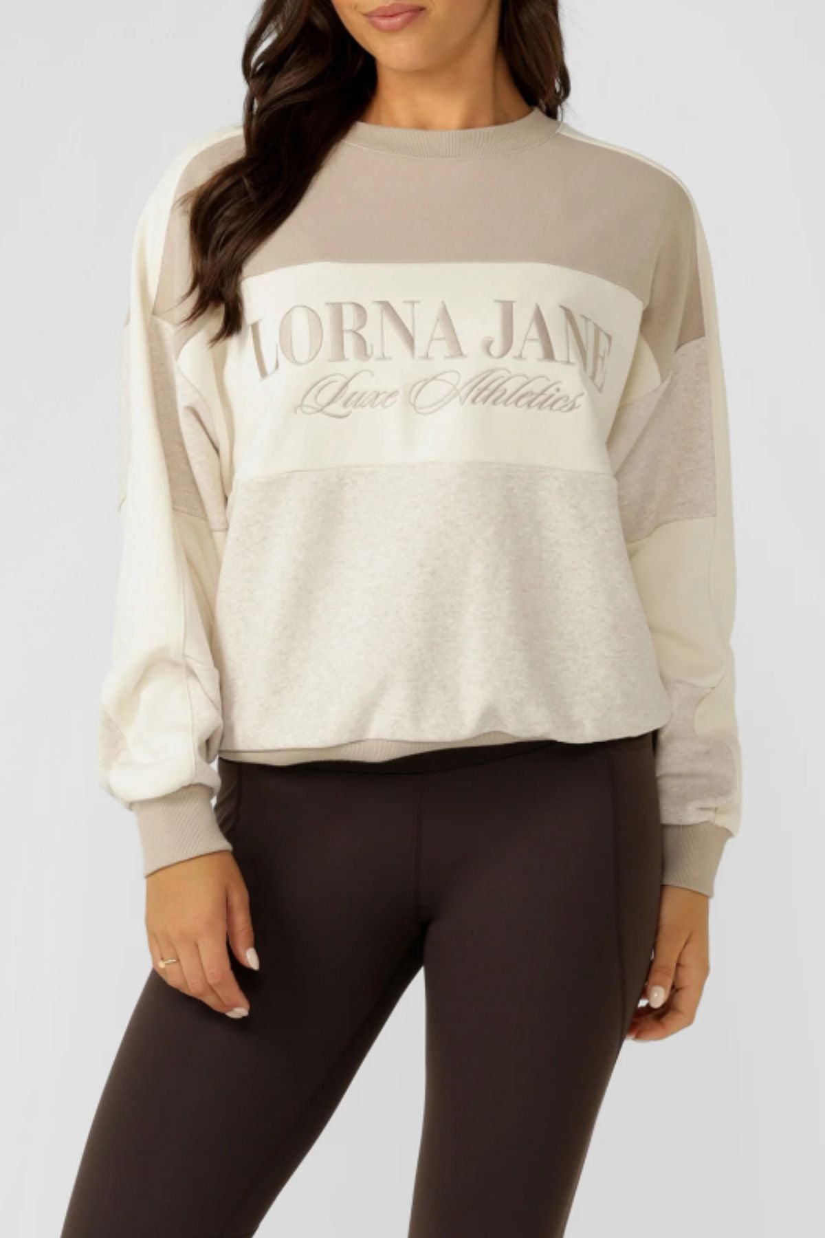 Model wears the Lorna Jane Lorna Luxe Panelled Sweat in light beige and cream cotton-blend terry with "LORNA JANE Luxe Athletics" front detail, paired with dark brown leggings, against a plain backdrop for a retro sport look.