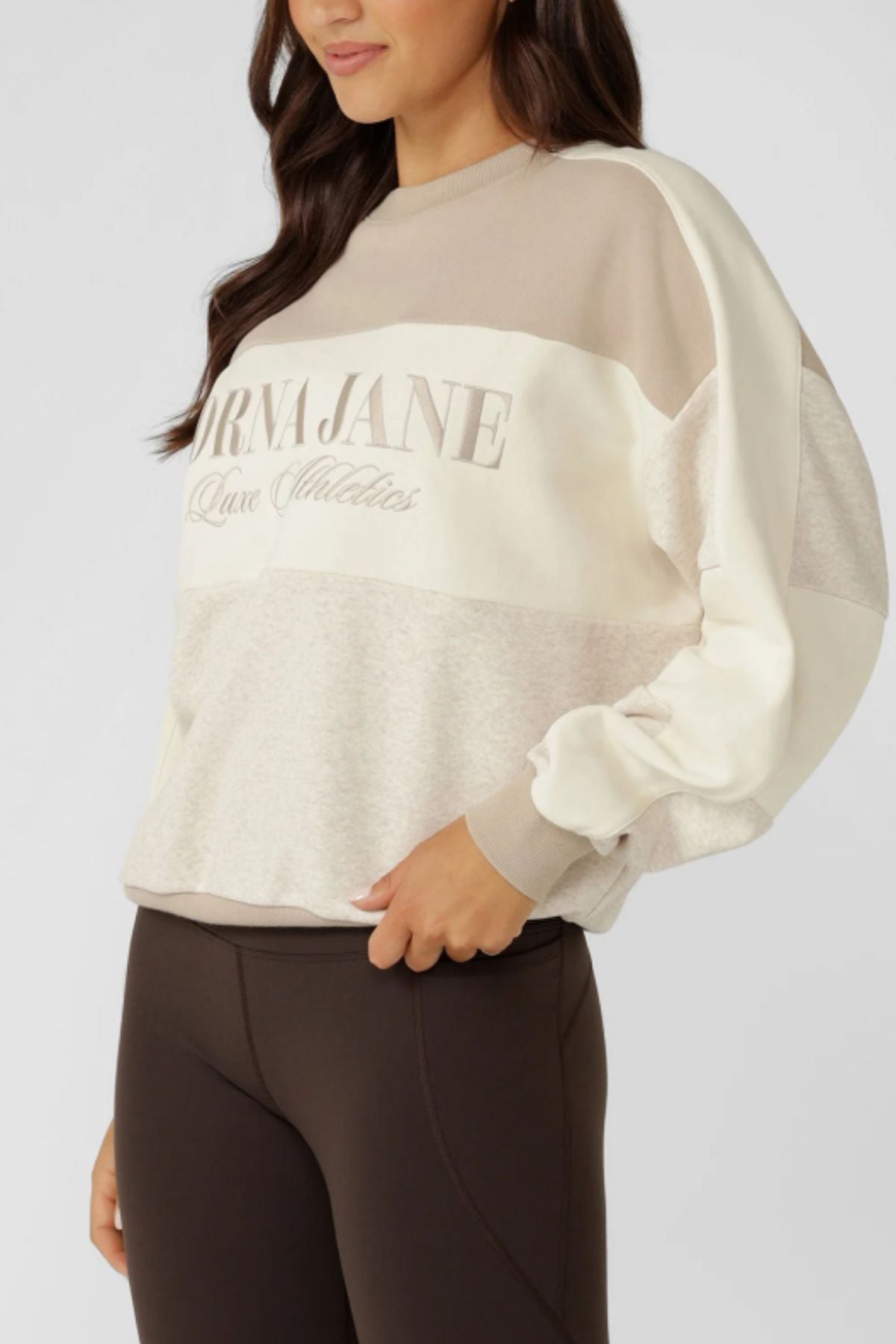 A woman wears the Lorna Jane Lorna Luxe Panelled Sweat, a loose, color-blocked cream and beige cotton-blend sweatshirt with an embroidered logo, paired with dark brown leggings for a retro sport look.