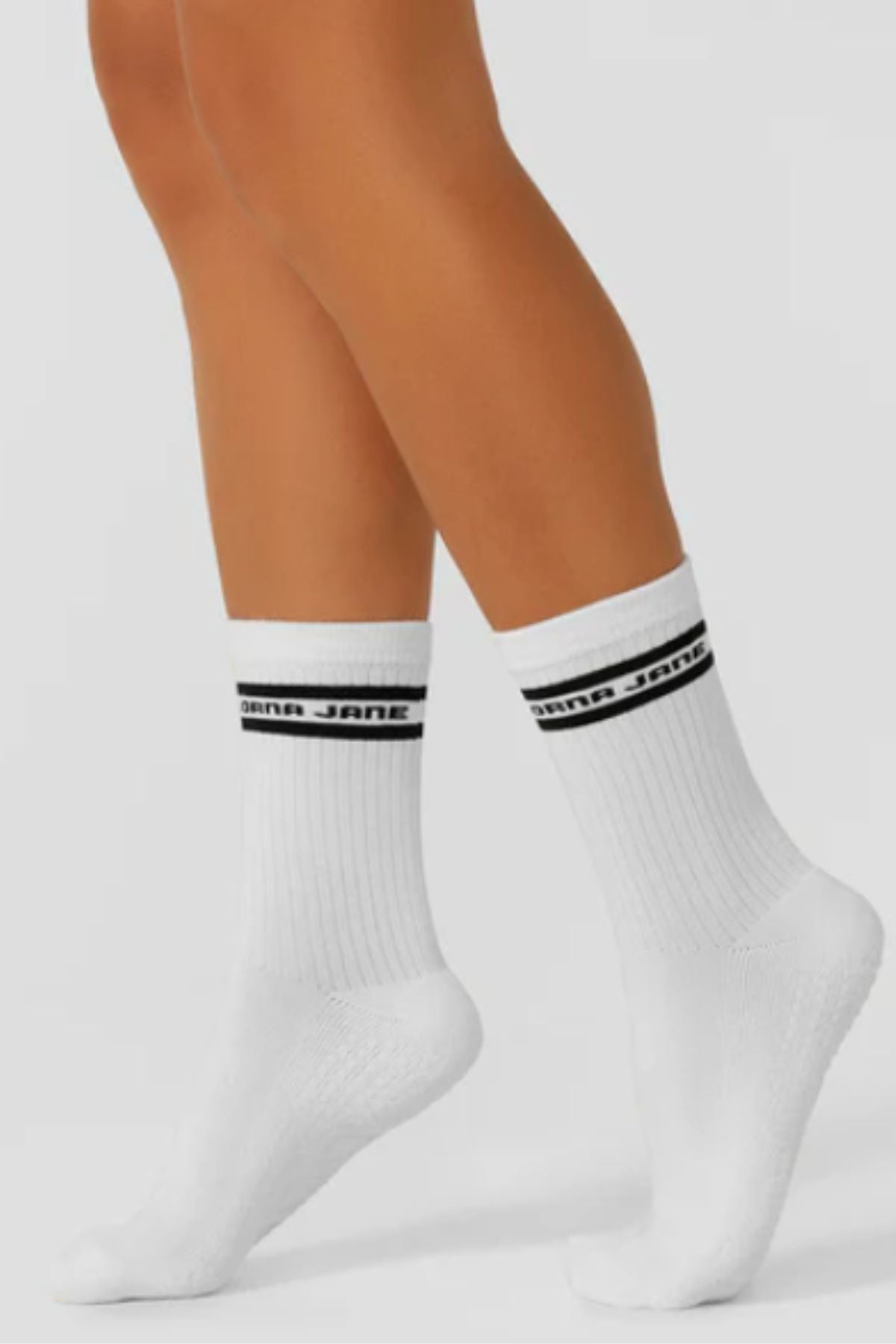 A person stands on tiptoe against a plain background, showcasing Lorna Jane Pilates Grip Crew Socks—white ribbed crew socks with two black stripes and "JANA JANE" text—offering stylish, non-slip comfort for everyday wear.