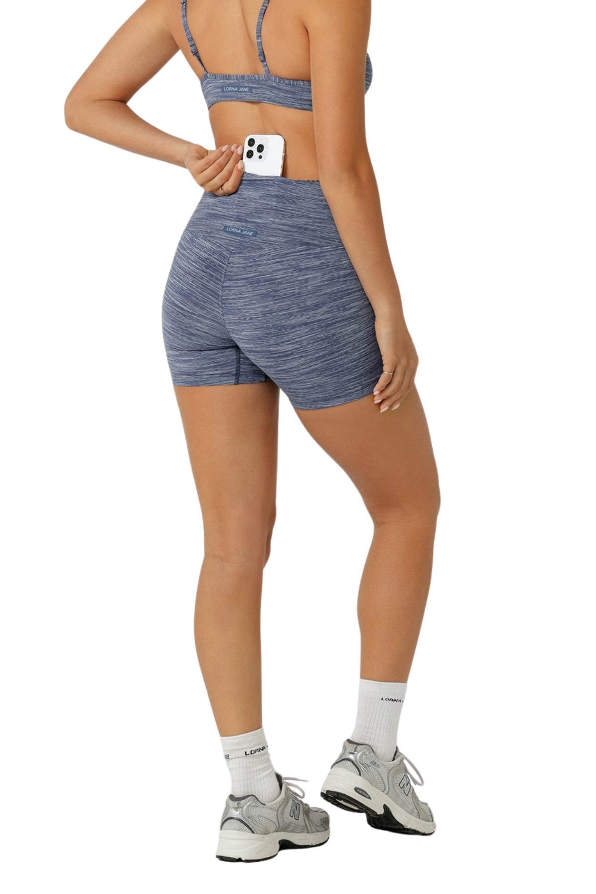 A person in blue Lorna Jane Retro 12cm Bike Shorts with a high-rise waistband places a smartphone in the back pocket. They wear white socks and gray sneakers, photographed from behind against a white background.