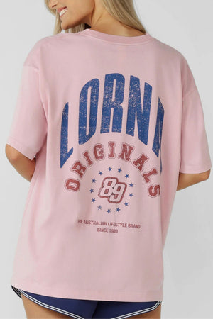 A woman with long blonde hair wears the Lorna Jane Revival Washed Boyfriend T-Shirt, featuring "Lorna Originals 89" in bold blue and red on the back. She is turned away, showing off the design, paired with navy blue shorts.