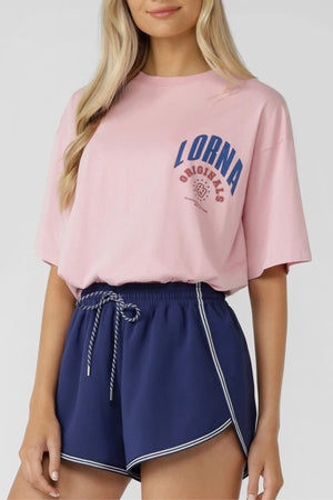 A woman wears the Lorna Jane Revival Washed Boyfriend T-Shirt in vintage pink with "LORNA ORIGINALS" blue curved lettering, paired with navy athletic shorts with white trim and a striped drawstring. 100% cotton for all-day comfort.