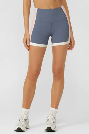 Someone wearing the Lorna Jane Set Point 2 Pocket 12cm Bike Short in blue and pink with a white band at the hem, paired with white socks and sneakers, stands against a plain light background.