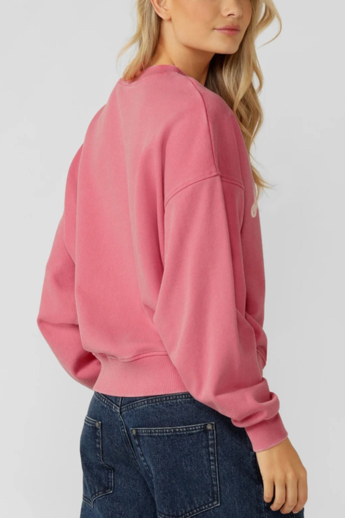 A woman with long blonde hair wears the Lorna Jane Sports Club Washed Sweat, a vintage-inspired oversized pink cotton terry sweatshirt, paired with dark blue jeans, standing sideways and looking over her shoulder against a plain backdrop.