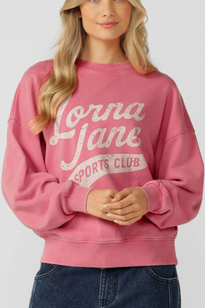 A woman with long blonde hair wears the Lorna Jane Sports Club Washed Sweat, an oversized pink vintage sweatshirt with "Lorna Jane Sports Club" in white text, styled with blue jeans against a plain light background.