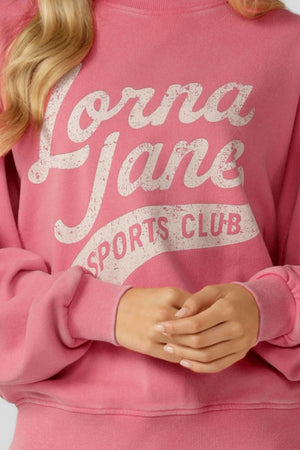 A person wearing the Lorna Jane Sports Club Washed Sweat, an oversized pink vintage sweatshirt with "Lorna Jane Sports Club" in white, shown from torso to hands with arms crossed at the waist.