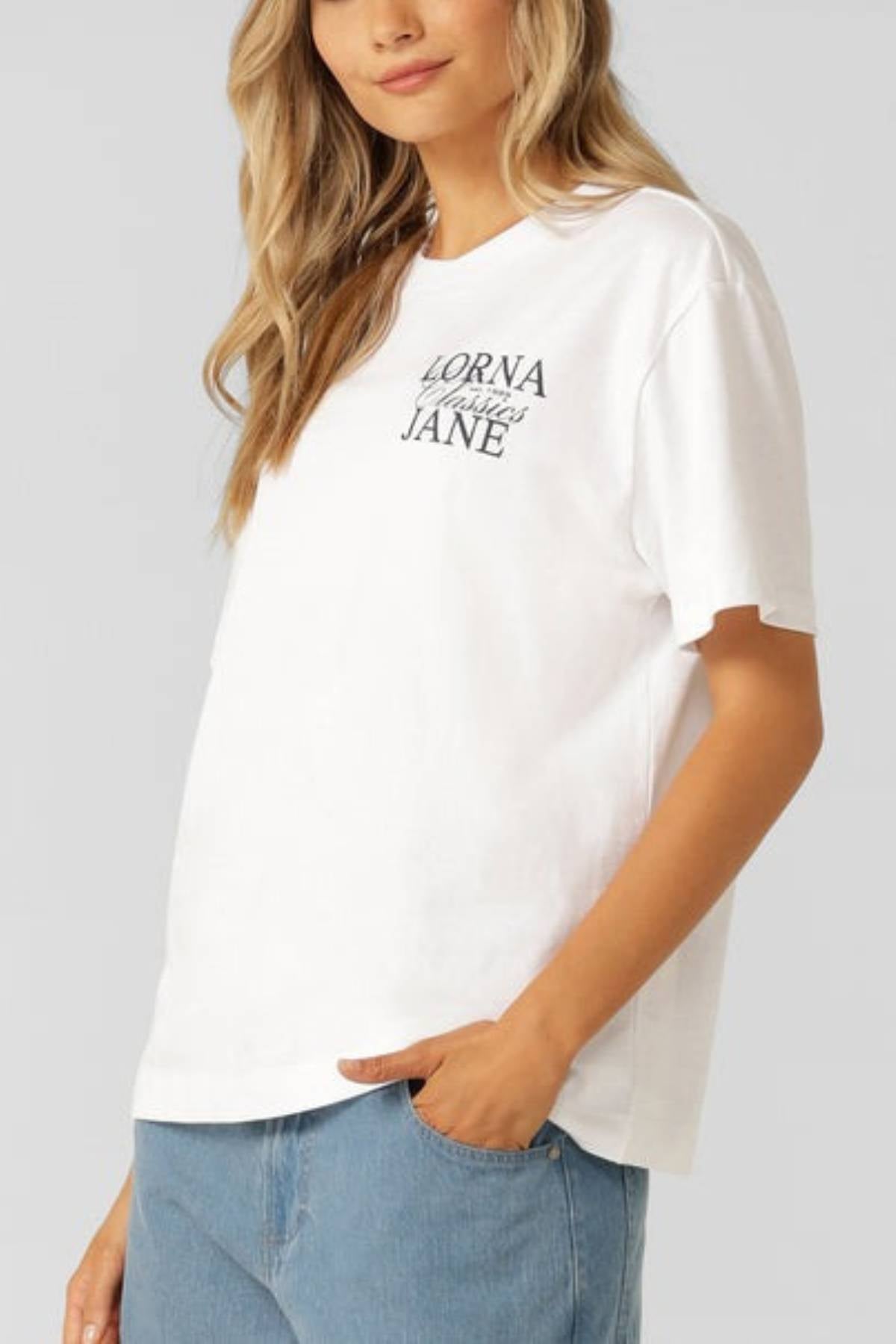 A woman with long, wavy blonde hair wears an oversized Lorna Jane Sprint Classic Fit T-Shirt in soft cotton jersey and light blue jeans, standing with one hand in her pocket against a plain light gray background.